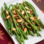 Top down view of a white rectangle serving plate with whole green beans with almonds and crispy shallots.